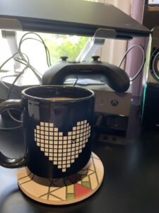 a coffee mug with an heart shape containing an Xbox logo, and behind it an Xbox development kit with a controller on top
